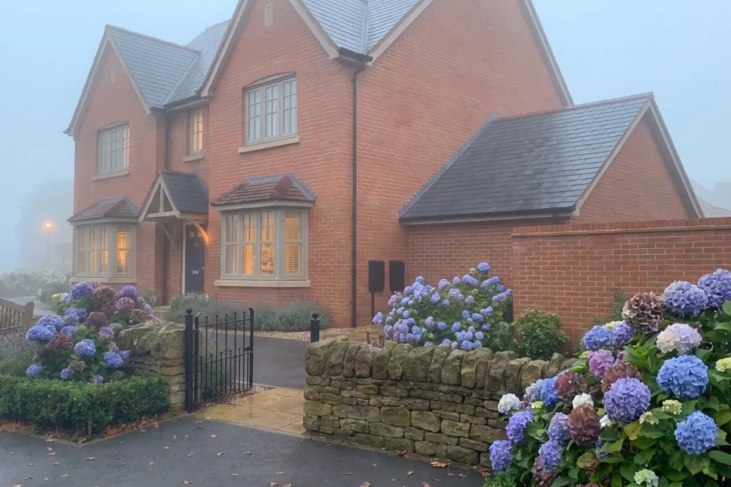 Two-story red brick house with a black front gate, stone walls, and blooming blue hydrangea bushes on a misty morning.