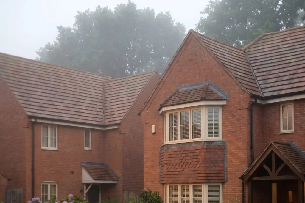 Multi-level rooflines with earthy brown clay tiles and prominent forward-facing gables on a red brick house exterior against a misty tree canopy.