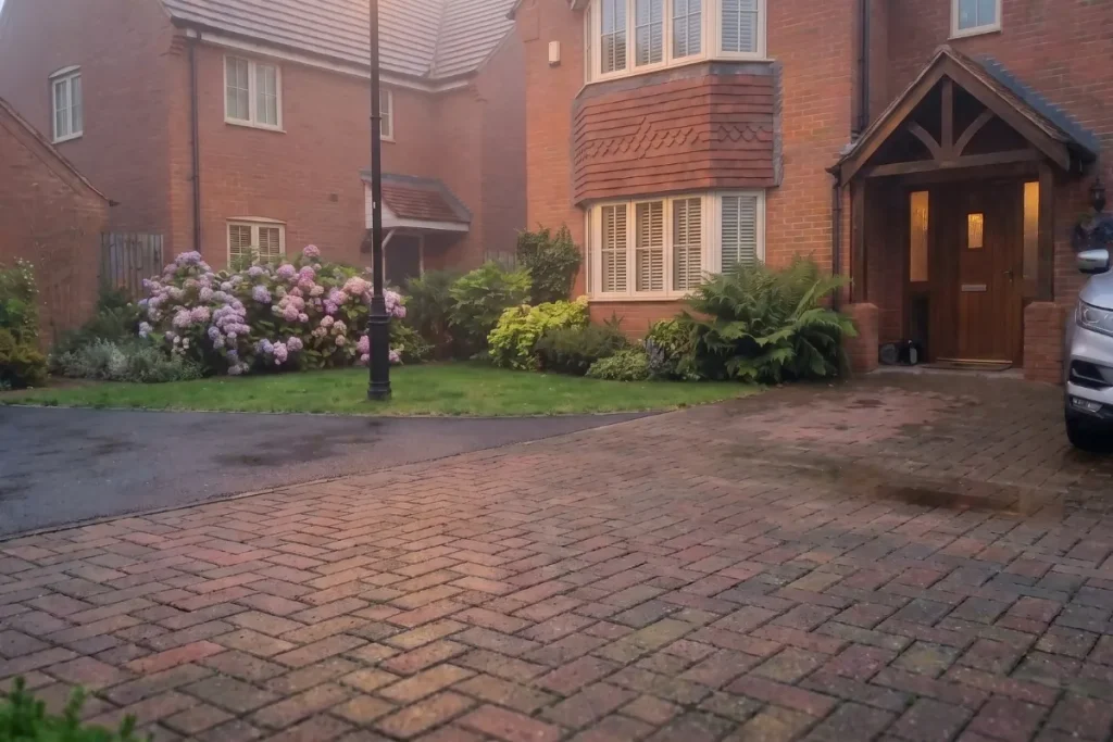 A block paved driveway sweeping across the front yard of a red brick house with a traditional timber entrance porch and a lawn border.