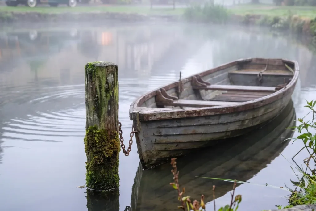 Close-up of an old wooden rowboat chained to a mossy wooden post in a misty pond, with ripples in the calm water.